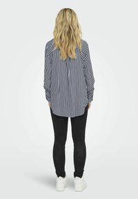 Camisa de manga larga a rayas en azul marino y blanco, abotonada por la parte de atrás, combinada con jeans skinny negros y zapatillas blancas. Diseño casual.