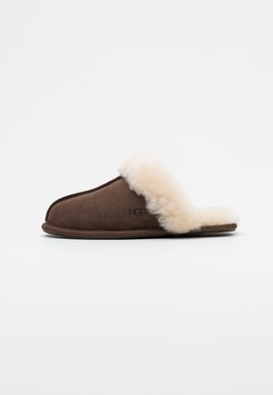 Brune semsket tøffel med en kremfarget saueskinnsfôring og rund tå. Har svart UGG-logo på siden og en flat såle.