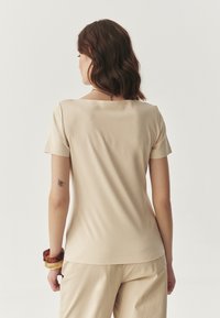 Beige top met korte mouwen en een vierkante halslijn. Gemaakt van soepele stof, heeft het een aansluitend ontwerp met een lichte rek en een ronde zoom.