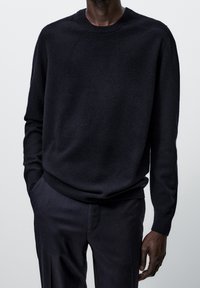 Pull en maille bleu marine avec un col rond et des poignets côtelés, confectionné dans un tissu doux. Porté avec un pantalon foncé assorti.