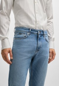 Light blue denim jeans with a straight-leg design, five pockets, and subtle stitching details. Paired with a white striped long-sleeve shirt.