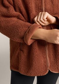 Brown, textured fleece jacket with a soft interior, featuring a smooth zippered front and rolled cuffs. Adjacent black leggings visible.