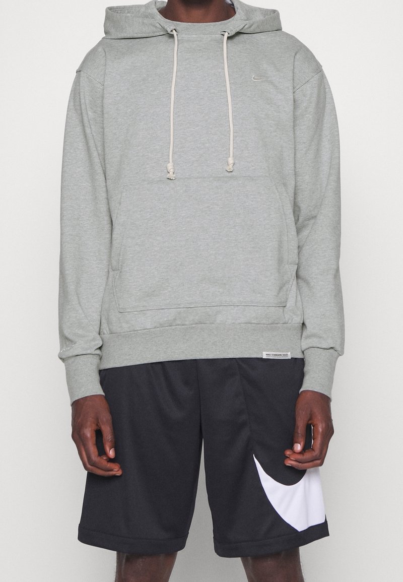 Personne portant un sweat à capuche gris clair avec une poche avant et un short de sport noir avec un grand logo Swoosh blanc sur une jambe.