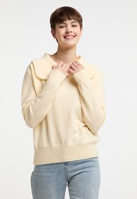 Cremefarbenes Sweater mit breitem Kragen, weichem Stoff, gerippten Bündchen und Saum. Das Modell trägt eng anliegende blaue Jeans. Neutraler Hintergrund.
