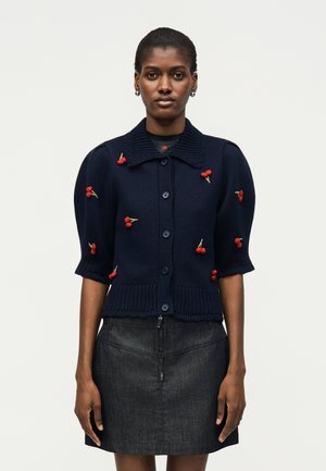 Γυναίκα που φοράει ένα σκούρο μπλε ζακέτα με κόκκινη κεντημένη κερασιά και μια σκοτεινή τζιν φούστα, στέκεται μπροστά σε ένα απλό φωτεινό φόντο.