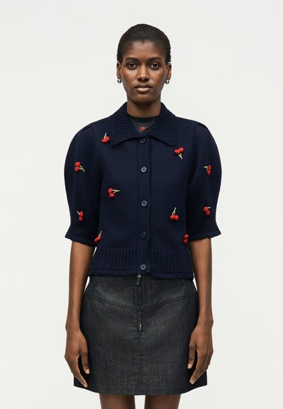 Kvinna bär en marinblå cardigan med röda körsbärsbroderier och en mörk denimkjol, stående mot en enkel ljus bakgrund.