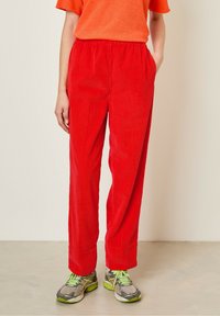 Pantalons en velours côtelé rouge avec une taille élastique, dotés de poches latérales et d'une coupe ample, associés à des chaussures de sport grises avec des accents verts.