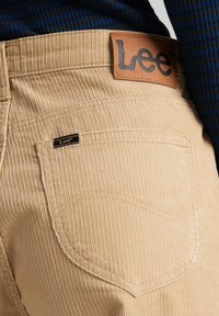 Beiga kordfladdriga byxor har en läderpatch med präglad "Lee"-logotyp och en liten svart "Lee"-tagg på bakfickan.