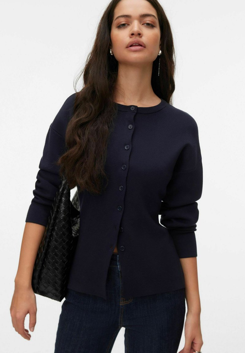 Cardigan bleu marine avec un col rond, une patte de boutonnage à l'avant et des manches longues. Présente une texture lisse et est associé à un sac à main noir tissé.