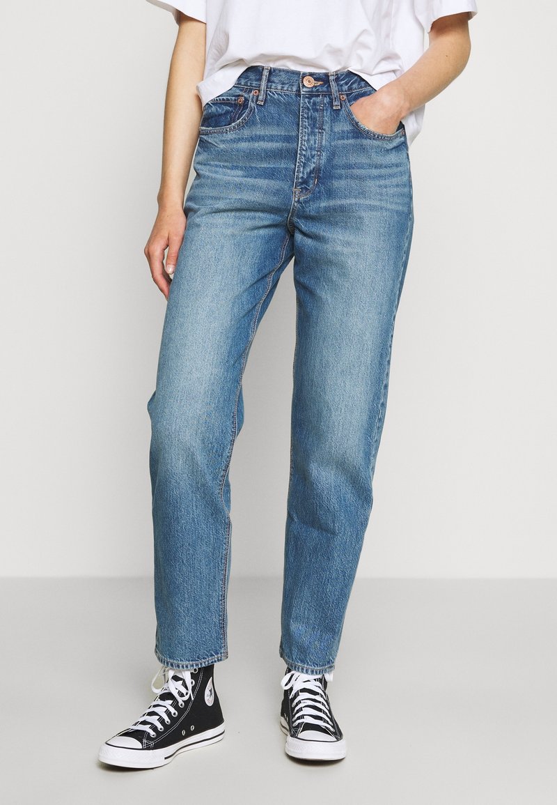 Personne portant un jean bleu taille haute, coupe droite, avec un t-shirt blanc plié rentré à l’intérieur et des baskets montantes noires à lacets blancs.