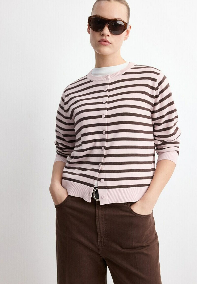 Femme portant des lunettes de soleil marron, un cardigan rayé à boutons rose clair et marron, un sous-vêtement blanc et un pantalon marron foncé les mains dans les poches.