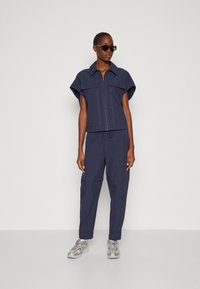 Marineblauw tweedelige outfit met een kortemouwen rits-top met twee borstzakken en tapered broek met een trekkoord in de taille en contrasterende stiksels.