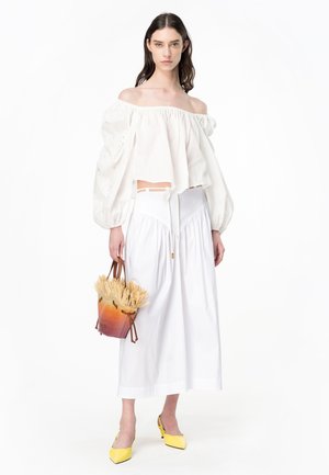Blusa bianca off-the-shoulder con maniche a sbuffo, abbinata a una gonna bianca alla caviglia. Scarpe gialle con tacco e borsa a tracolla con accenti in paglia.
