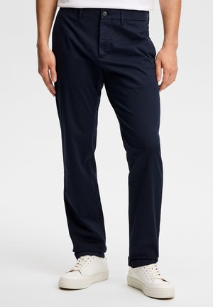 Mężczyzna w granatowych spodniach o dopasowanym kroju i białych sneakersach, stojący na jednolitym tle.