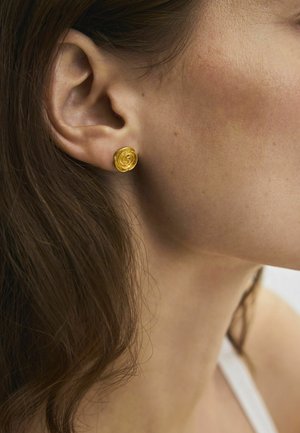 Nahaufnahme des Ohrs einer Frau mit langen braunen Haaren, die einen kleinen, runden goldenen Ohrring in Form einer Blume trägt.