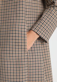 Brązowo-czarna tkanina w kratę o gładkiej fakturze. Wzór obejmuje kieszeń z boku oraz długi rękaw.