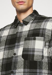 Gesteppte Knopfjacke in Schwarz, Weiß und grauem Karomuster. Mit umklappbarem Kragen und einer Brusttasche mit Etikett. Texturiertes Material.