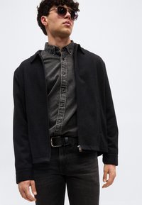 Άνδρας με σκούρα γυαλιά ηλίου, μαύρο μπουφάν με φερμουάρ πάνω από ξεθωριασμένο μαύρο πουκάμισο με κουμπιά, και μαύρο τζιν μπροστά σε απλό φόντο.