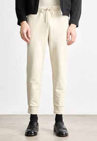 Joggery w jasnobeżowym kolorze wykonane z miękkiego materiału, ze sznurkiem w talii. Dopasowany krój zwężany ku dołowi, z mankietami przy kostkach, zestawione z czarnymi butami i skarpetkami.