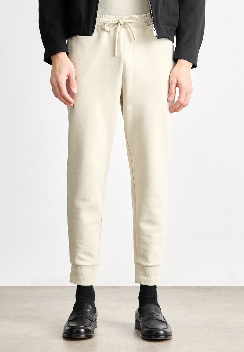 Joggery w jasnobeżowym kolorze wykonane z miękkiego materiału, ze sznurkiem w talii. Dopasowany krój zwężany ku dołowi, z mankietami przy kostkach, zestawione z czarnymi butami i skarpetkami.