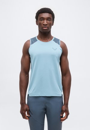 Lihaseline mees seisab, kandes helesinist varrukateta sporditoppi tumeõlgade ja sobivate pükstega valgel taustal.