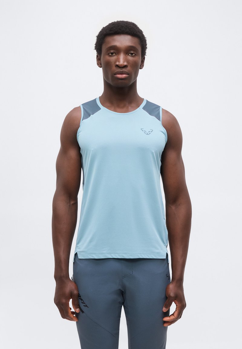 Μυώδης άνδρας όρθιος, φορά αμάνικη αθλητική μπλούζα ανοιχτού γαλάζιου χρώματος με σκούρες ωμοπλάτες και παντελόνι στο ίδιο χρώμα, σε λευκό φόντο.