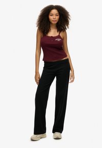 Bordeaux kleurige, aansluitende tanktop met kantafwerking en knoopdetail, gecombineerd met losse zwarte broeken. Model staat op een neutrale achtergrond.