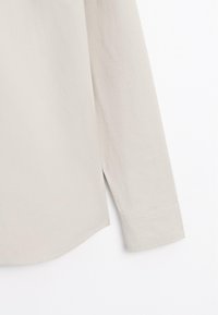 Chemise beige clair à manches longues avec un ourlet droit et une fente latérale, montrant le détail de la manche inférieure et du poignet sur un fond blanc.
