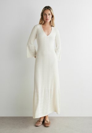 Kvinde står iført en lang hvid strikket kjole med bell-ærmer og V-hals, kombineret med beige flade snøresko mod en ensfarvet væg.