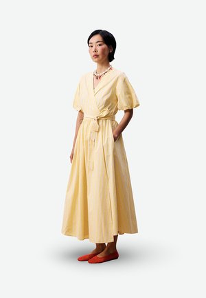 Femme debout portant une robe portefeuille jaune clair à manches bouffantes et des ballerines orange, les mains dans les poches de la robe, fond neutre.