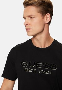 Camiseta de algodón negra con un logo en relieve y brillante que dice "GUESS EST. 1981" de forma destacada en el pecho.