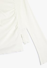 Top bianco a maniche lunghe con trama a costine, caratterizzato da dettaglio arricciato sul lato sinistro, orlo dritto e spacco laterale per un design aggiuntivo. Texture morbida.
