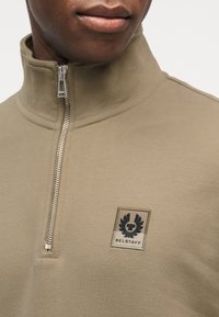 Gros plan d'une personne portant un haut zippé beige avec une fermeture éclair argentée et un logo Belstaff noir sur la poitrine gauche.