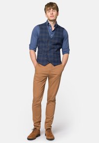 Granatowa kamizelka w kratę na niebieskiej koszuli, beżowe dopasowane spodnie oraz brązowe zamszowe buty. Kamizelka ma podwójne zapięcie i teksturowaną tkaninę.