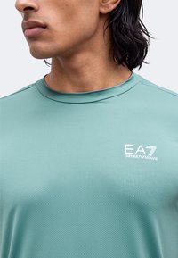 Άνδρας που φοράει ανοιχτό τιρκουάζ πουκάμι EA7 Emporio Armani με υφή μικρού μήκους και στρογγυλή λαιμόκοψη.
