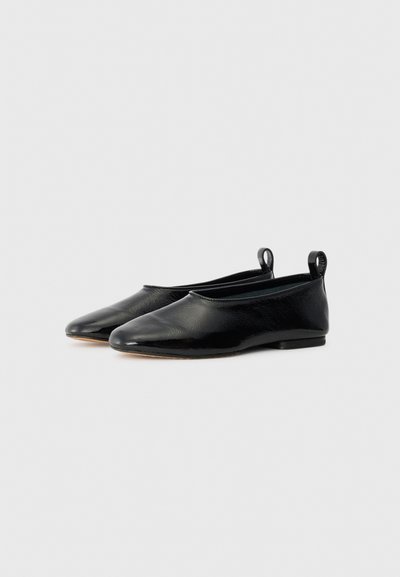 Ballerines noires en cuir avec une finition lisse, un bout pointu et un petit talon. Présentent de petites boucles au niveau du talon pour un enfilage facile.