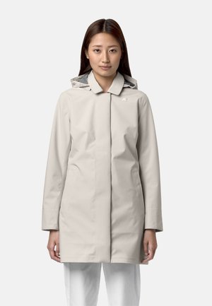 Młoda kobieta w beżowej kurtce przeciwdeszczowej z kapturem i białych spodniach, stojąca zwrócona przodem na tle jednolitego jasnego tła.