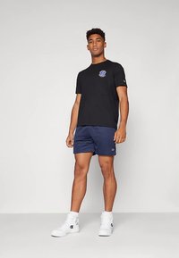 Juoda trumpomis rankovėmis medvilninė marškinėliai su grafiniu logotipu ant krūtinės, derinami su tamsiai mėlynais šortais ir baltais aukštais sportiniais bateliais.