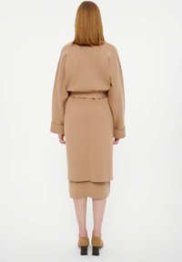 Cappotto di lunghezza al ginocchio, di colore marrone chiaro, con vita con cintura, maniche ampie e texture liscia, indossato sopra una gonna coordinata di lunghezza al ginocchio e scarpe con tacco.