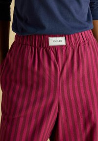 Ružové pásikavé šortky s elastickým pásom, s bielou etiketou s nápisom "JOULES" v čiernom texte. Mäkko tkanina a uvoľnený strih.