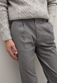 Pantaloni di lana grigia con una vestibilità sartoriale, caratterizzati da pieghe frontali e una texture sottile. Sopra viene indossato un maglione lavorato a maglia di colore grigio chiaro.
