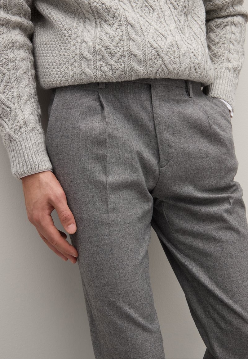 Pantaloni di lana grigia con una vestibilità sartoriale, caratterizzati da pieghe frontali e una texture sottile. Sopra viene indossato un maglione lavorato a maglia di colore grigio chiaro.