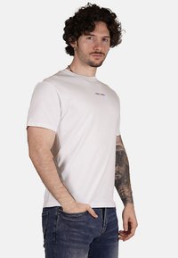 T-shirt bianco a maniche corte con collo rotondo, caratterizzato da un piccolo logo multicolore sul petto. Materiale in cotone con una texture liscia.