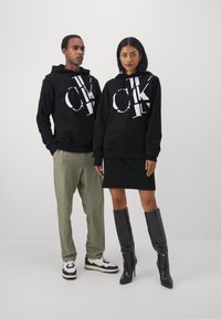 Čierne mikiny s veľkým bielym logom "CK", sivé nohavice, čierna sukňa a čierne vysoké čižmy. Hladká tkanina, voľný strih, minimalistický dizajn.