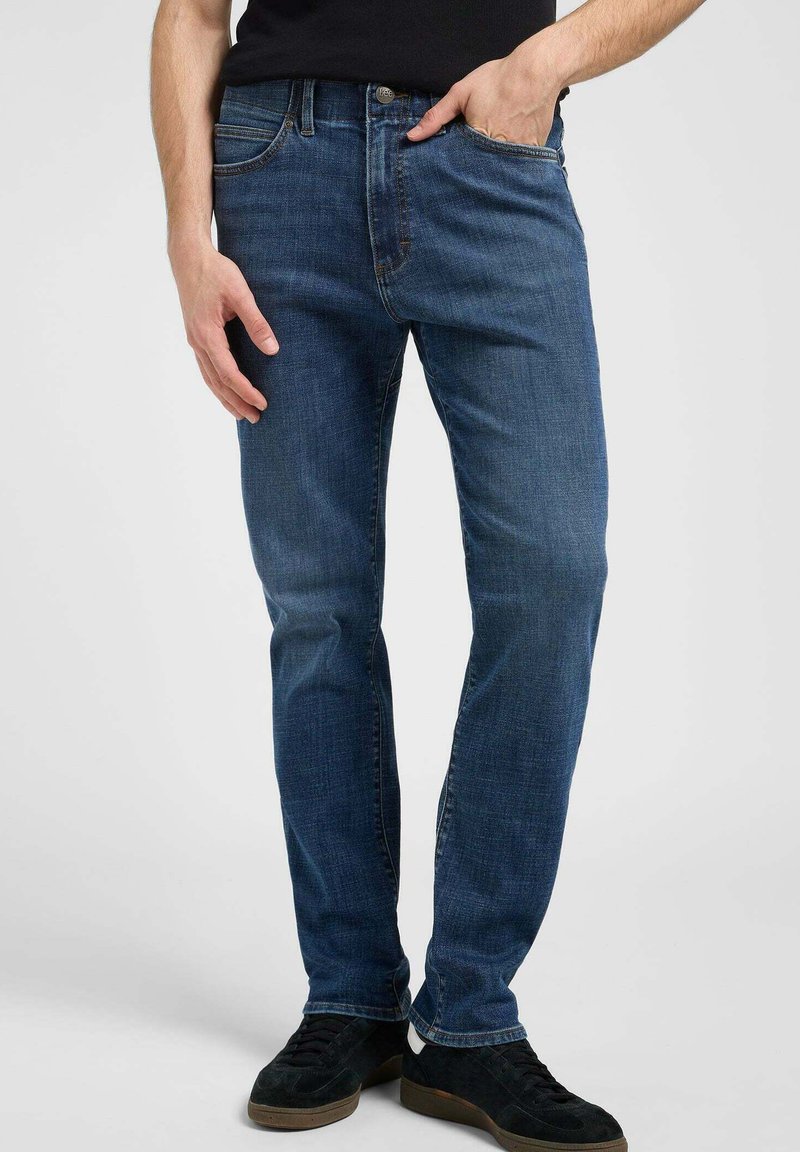 Vaqueros de mezclilla azul oscuro con un corte ajustado, diseño de cinco bolsillos y sutiles acentos de costura. Llevados con zapatillas negras.