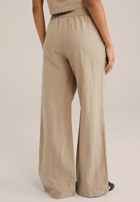 Pantaloni larghi beige realizzati in un tessuto texture con elastico in vita. Orlo parzialmente infilato con spacchi laterali. Calzature nere visibili.