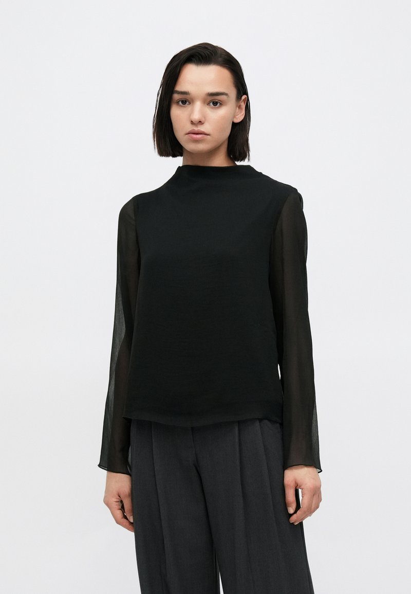 Μαύρο μακρυμάνικο τοπ με υψηλό λαιμό και διαφανή μανίκια, σετ με γκρι παντελόνια φαρδιάς γραμμής, με ομαλή υφή.