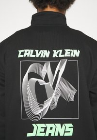 Dos d'une veste noire avec le texte vert "Calvin Klein Jeans" et un logo "CK" stylisé blanc dans un cadre carré.