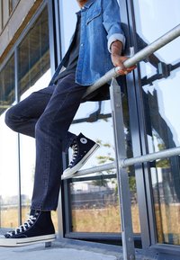 Jeansjacke mit aufgerollten Ärmeln über einem schwarzen Hemd, kombiniert mit dunklen Jeans und schwarzen High-Top-Converse-Sneakern auf einem Geländer.