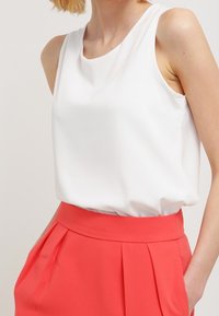 White sleeveless top with a smooth texture and rounded neckline, paired with bright coral pleated high-waisted pants.